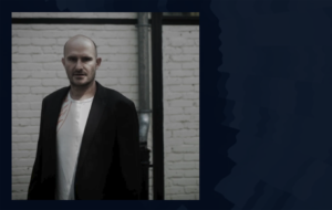 Un homme chauve portant un blazer sombre et une chemise blanche se tient devant un mur de briques blanches faiblement éclairé, affichant un comportement d'entreprise sérieux alors qu'il regarde la caméra. Photographe Xavier Granet