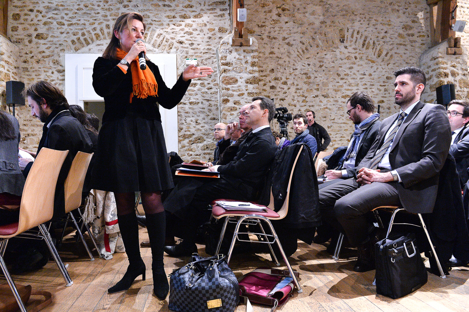 Une femme se tient debout et parle dans un microphone lors d'une conférence, tandis que les participants assis en rangs écoutent attentivement dans une salle rustique aux murs de pierre. Des sacs et des manteaux sont posés sur le sol près de leurs chaises. Photographe Xavier Granet
