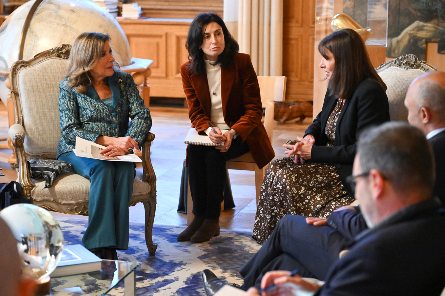 Trois femmes sont assises dans une pièce formelle, engagées dans une conversation, avec des cahiers à la main. Deux hommes avec des cahiers sont visibles au premier plan, partiellement flous. Des étagères et un grand globe terrestre sont visibles à l'arrière-plan. Photographe Xavier Granet