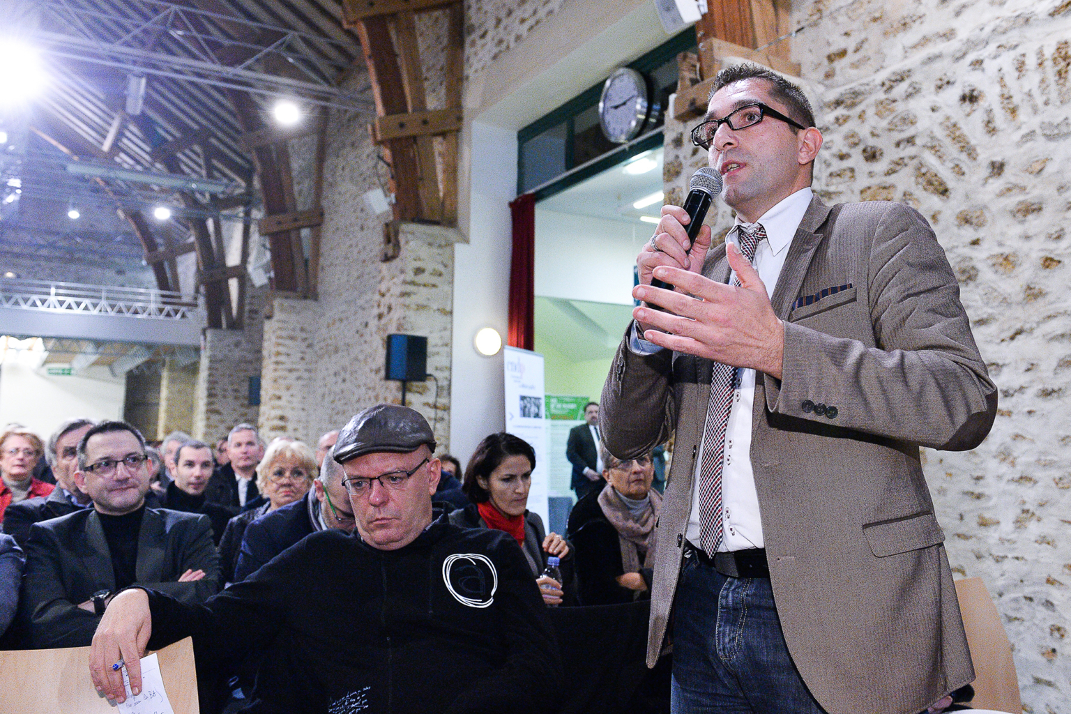 Un homme vêtu d'un blazer beige parle dans un microphone lors d'un événement en intérieur, tandis qu'un public l'écoute attentivement. Les murs de la salle sont en pierre et les poutres en bois. Plusieurs personnes sont assises, certaines regardant vers l'orateur. Photographe Xavier Granet