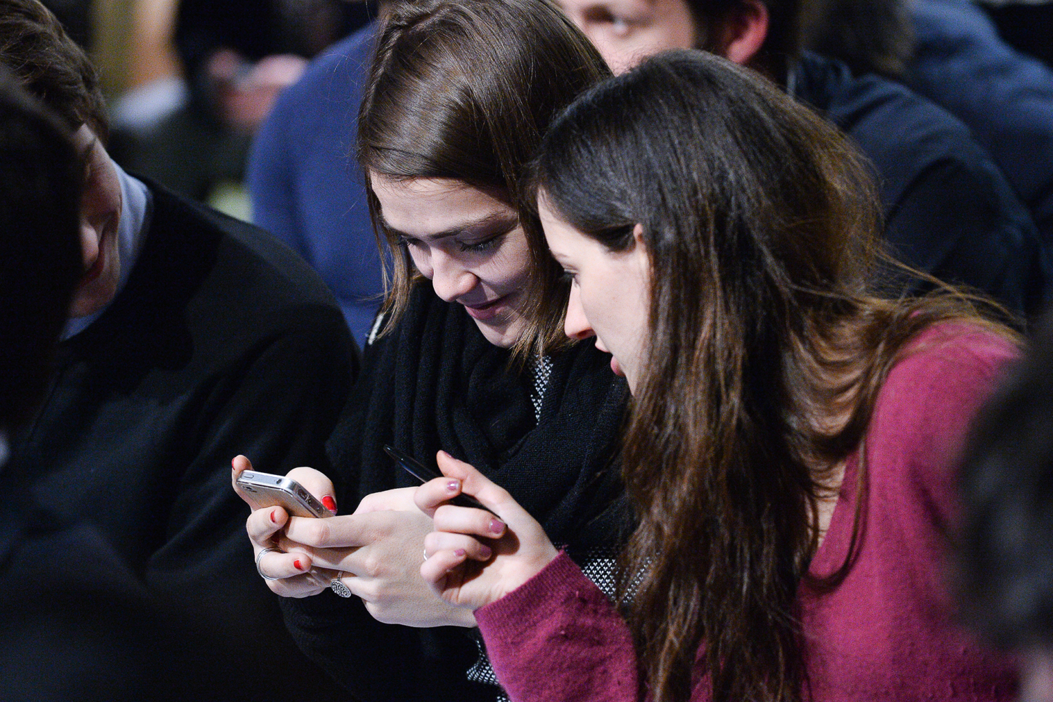 Deux femmes assises l'une à côté de l'autre dans une foule, regardant toutes deux attentivement un smartphone que l'une d'entre elles tient, semblant engagées et concentrées sur l'écran. Photographe Xavier Granet