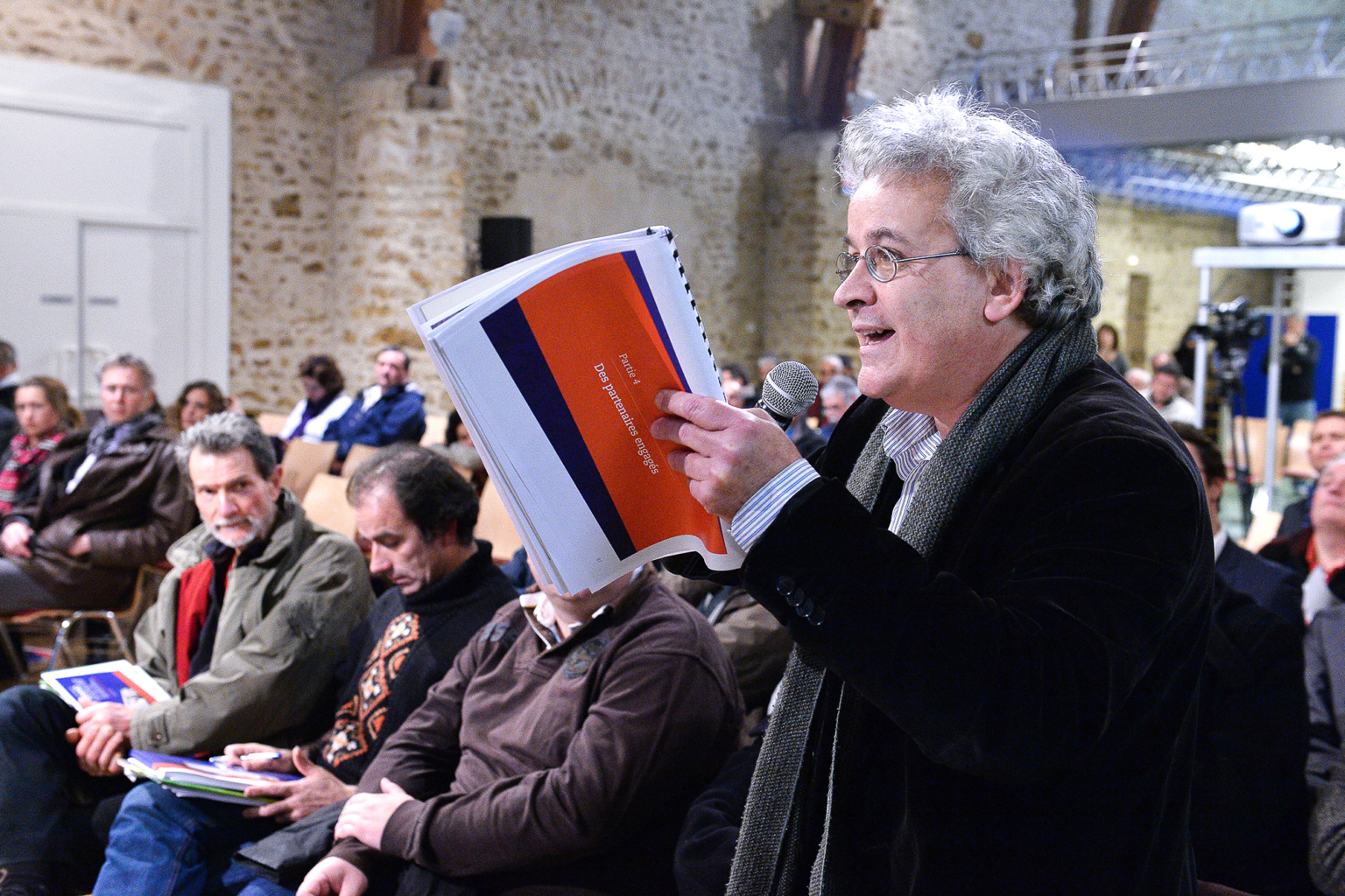 Un homme tenant un micro et un document s'adresse à un public assis dans une salle aux murs de pierre. D'autres personnes écoutent attentivement en tenant des documents similaires. Photographe Xavier Granet