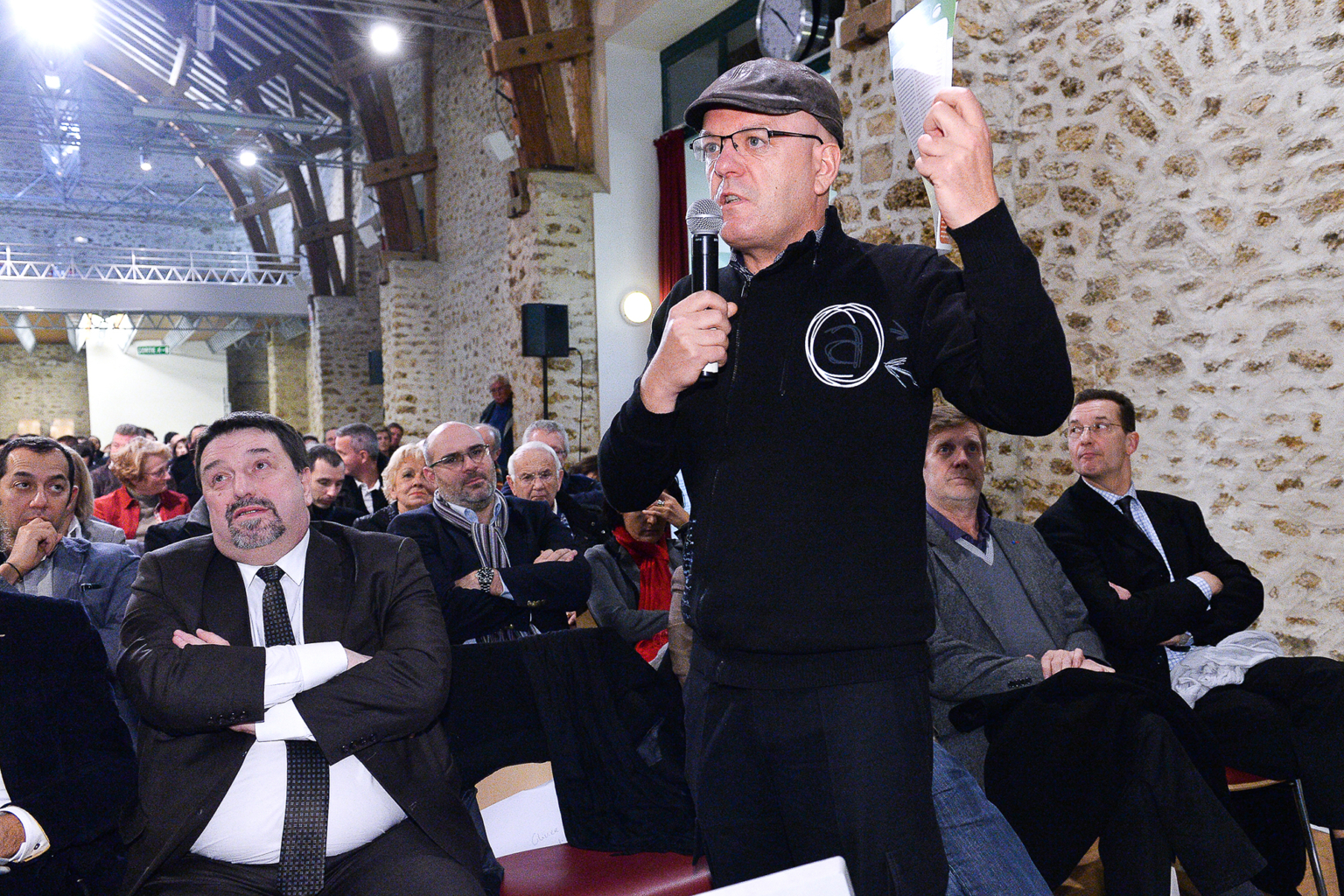 Un homme vêtu d'un pull noir et d'une casquette parle dans un micro, debout au milieu d'un public assis, dans une salle intérieure rustique aux murs de pierre et aux poutres en bois. Les gens écoutent attentivement, certains les bras croisés. Photographe Xavier Granet