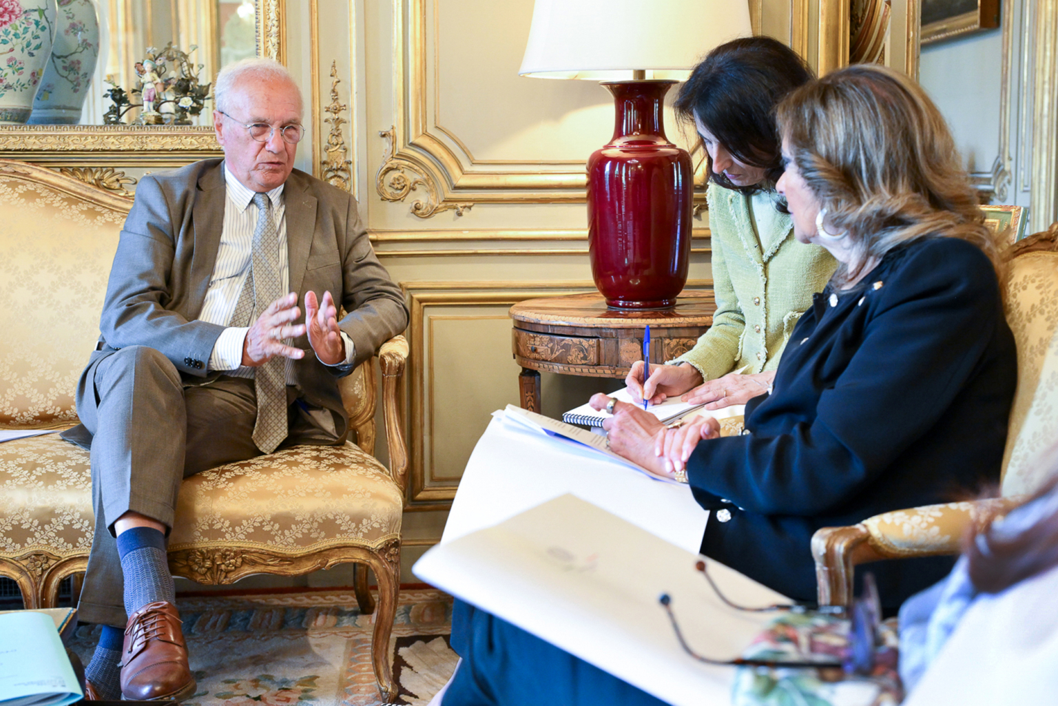 Un homme en costume est assis sur un canapé à motifs dorés. Il parle et fait des gestes avec ses mains. Deux femmes sont assises en face de lui, prenant des notes, tandis qu'une autre femme écoute attentivement dans une salle élégante au décor orné. Photographe Xavier Granet