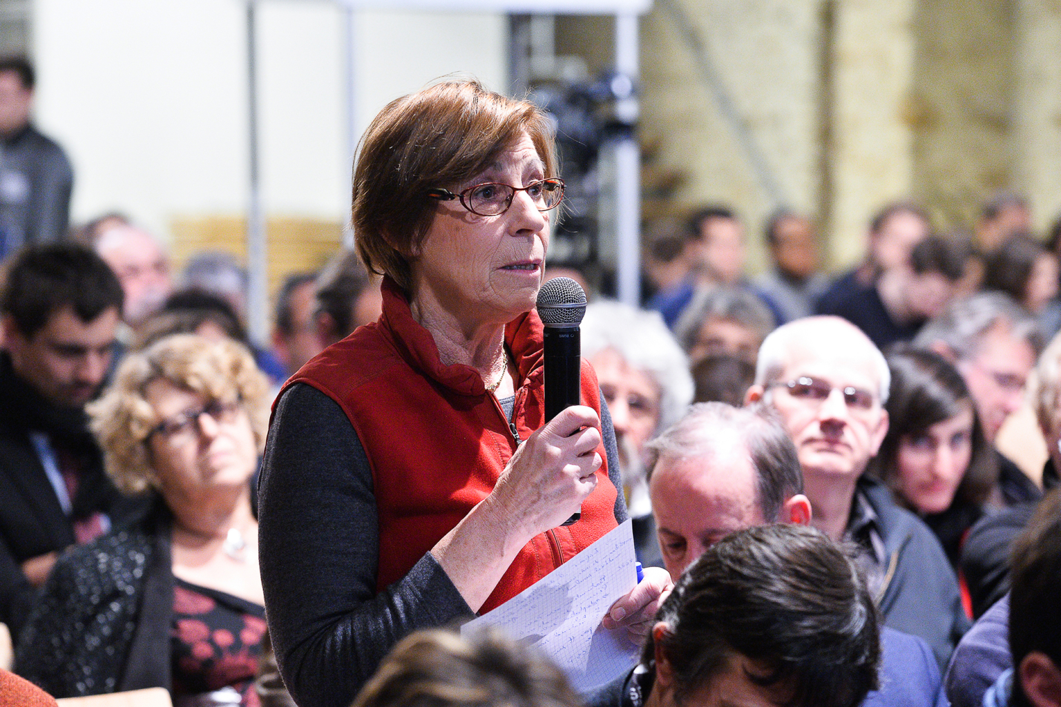 Une femme âgée portant des lunettes et un gilet rouge tient un micro et parle debout dans une salle bondée de personnes assises, dont beaucoup écoutent attentivement. Photographe Xavier Granet