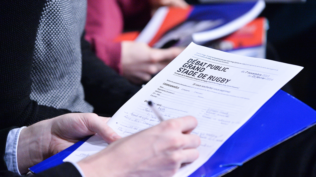 Une personne tient un dossier bleu et remplit au stylo un formulaire intitulé "Débat public Grand Stade de Rugby", assise parmi d'autres personnes, dont certaines tiennent des documents. Photographe Xavier Granet