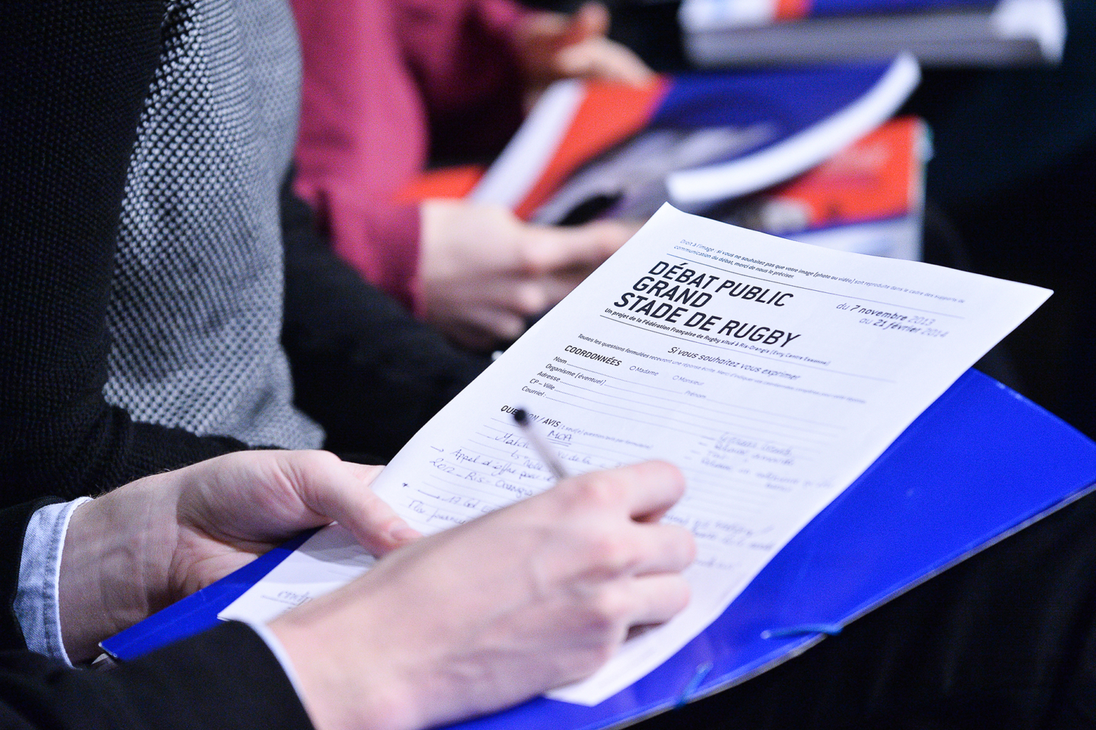 Une personne tient un dossier bleu et remplit au stylo un formulaire intitulé "Débat public Grand Stade de Rugby", assise parmi d'autres personnes, dont certaines tiennent des documents. Photographe Xavier Granet