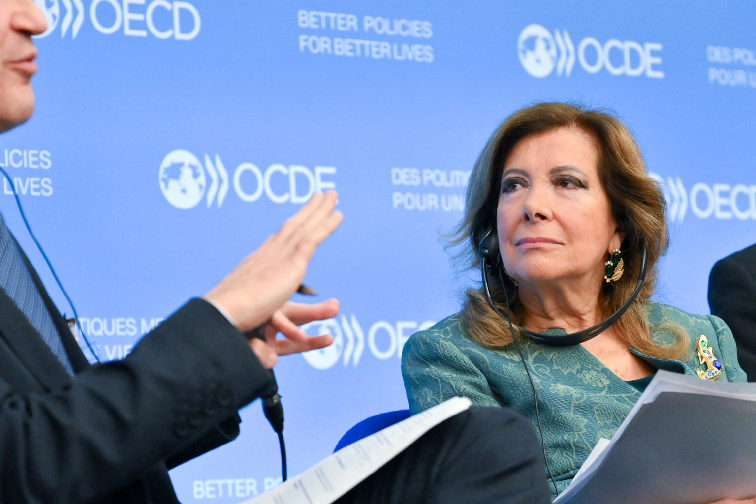 Une femme tenant des documents écoute attentivement un homme faisant un geste de la main lors d'un événement, avec un fond de scène de l'OCDE et le slogan "De meilleures politiques pour de meilleures vies" visible derrière eux. Photographe Xavier Granet