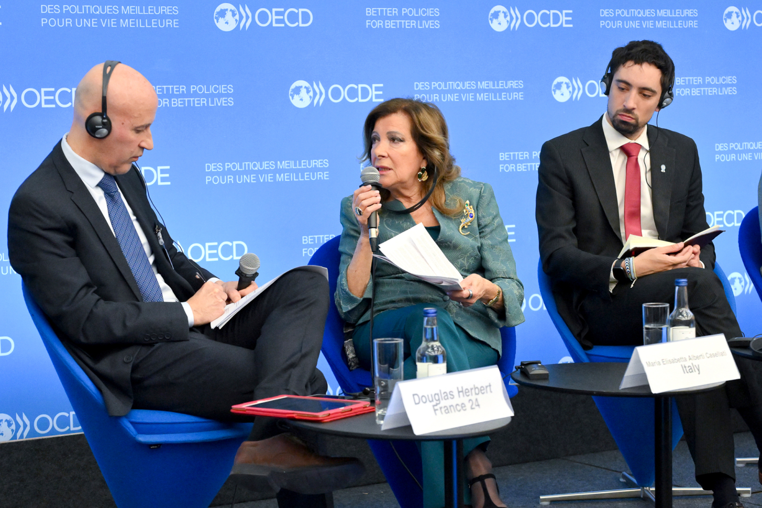 Trois personnes en tenue de soirée participent à un panel lors d'un événement de l'OCDE. La femme au centre parle dans un microphone, tandis que les deux hommes de chaque côté écoutent. Des plaques nominatives et un fond bleu de l'OCDE sont visibles derrière eux. Photographe Xavier Granet