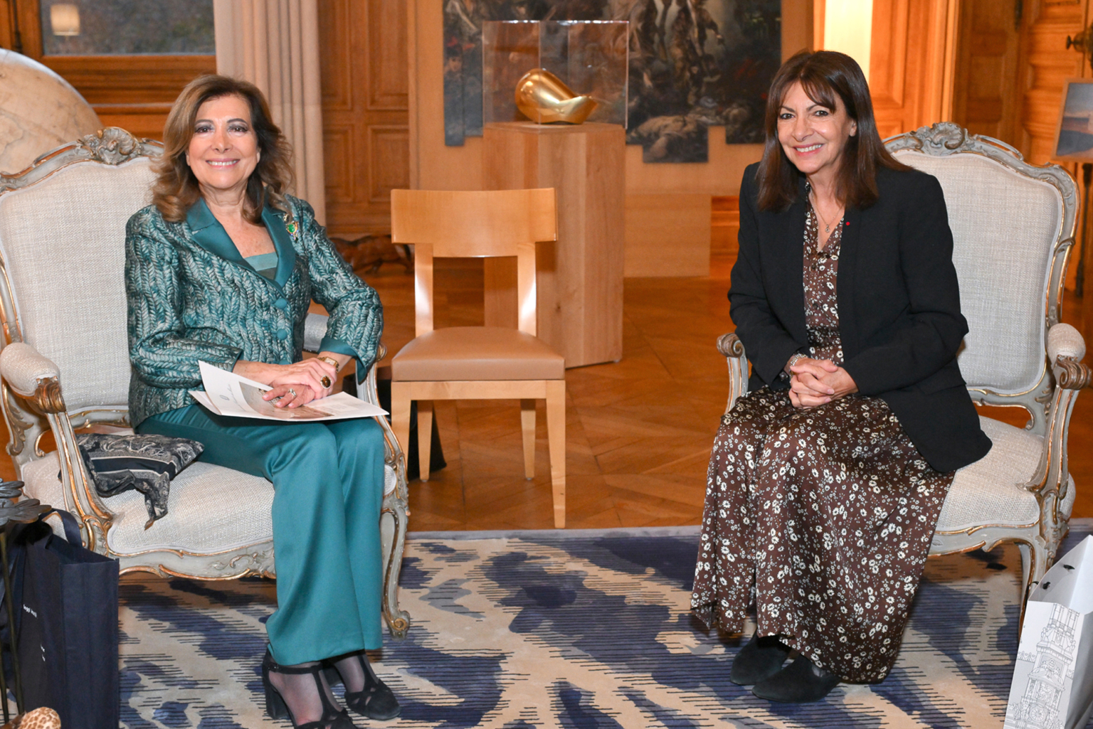 Deux femmes sont assises sur des chaises ornées dans une pièce chaudement éclairée, avec des murs en bois, un tapis à motifs et des œuvres d'art en arrière-plan. Toutes deux sourient ; l'une porte un tailleur vert, l'autre une robe à motifs et une veste noire. Photographe Xavier Granet