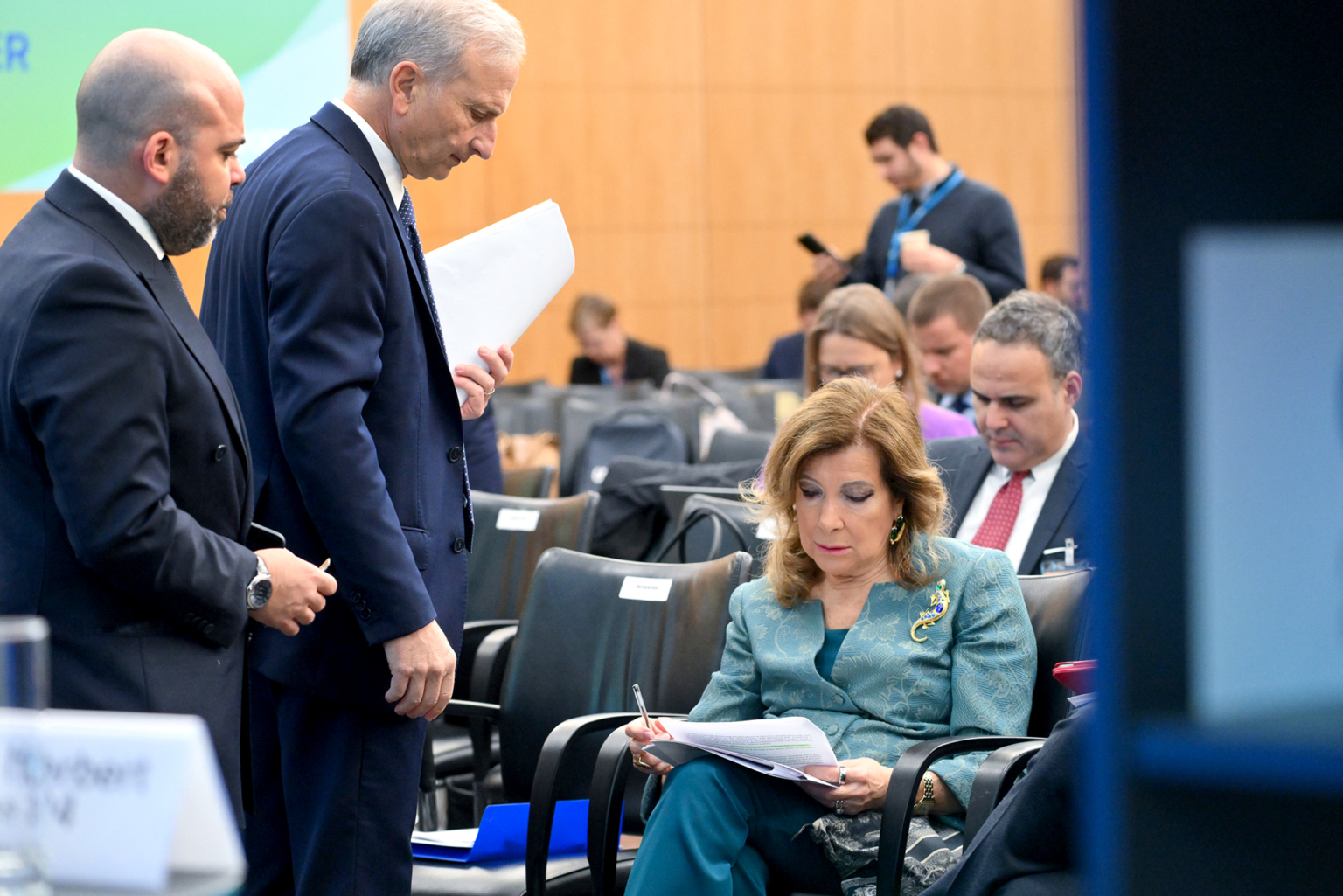 Une femme est assise et lit des documents dans une salle de conférence, tandis que deux hommes se tiennent à côté d'elle et discutent. D'autres participants sont assis à l'arrière-plan, certains lisant ou discutant également. L'atmosphère est formelle et professionnelle. Photographe Xavier Granet
