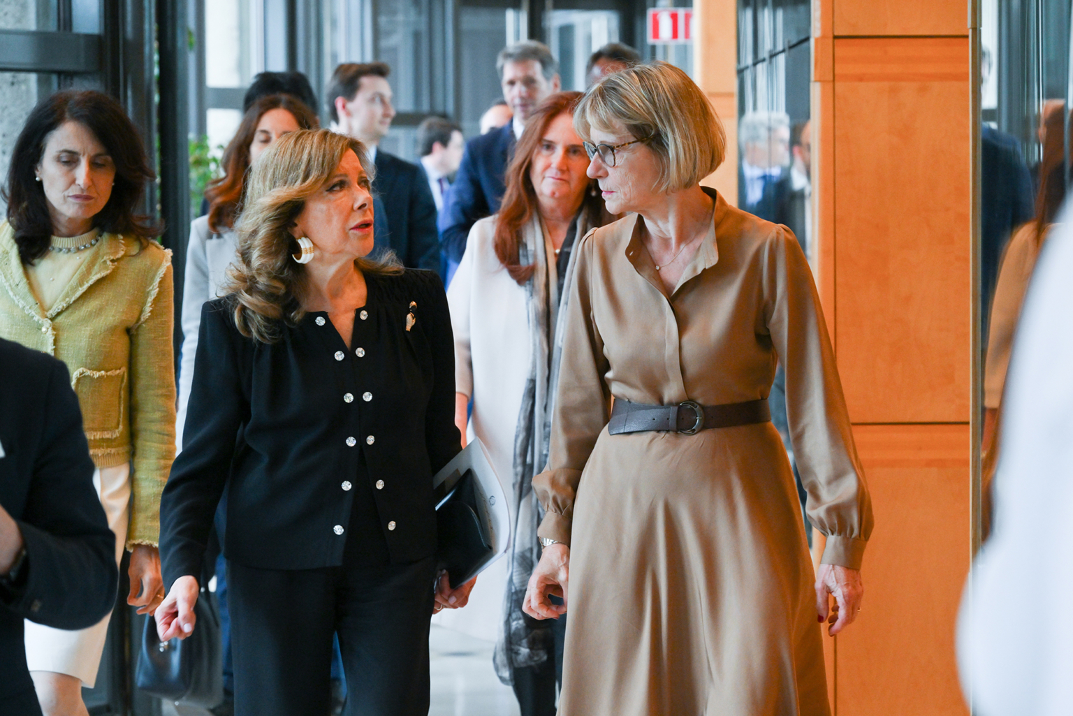 Un groupe d'adultes habillés professionnellement se promène à l'intérieur. Deux femmes à l'avant, l'une en tailleur noir et l'autre en robe beige, discutent tandis que d'autres les suivent. Photographe Xavier Granet