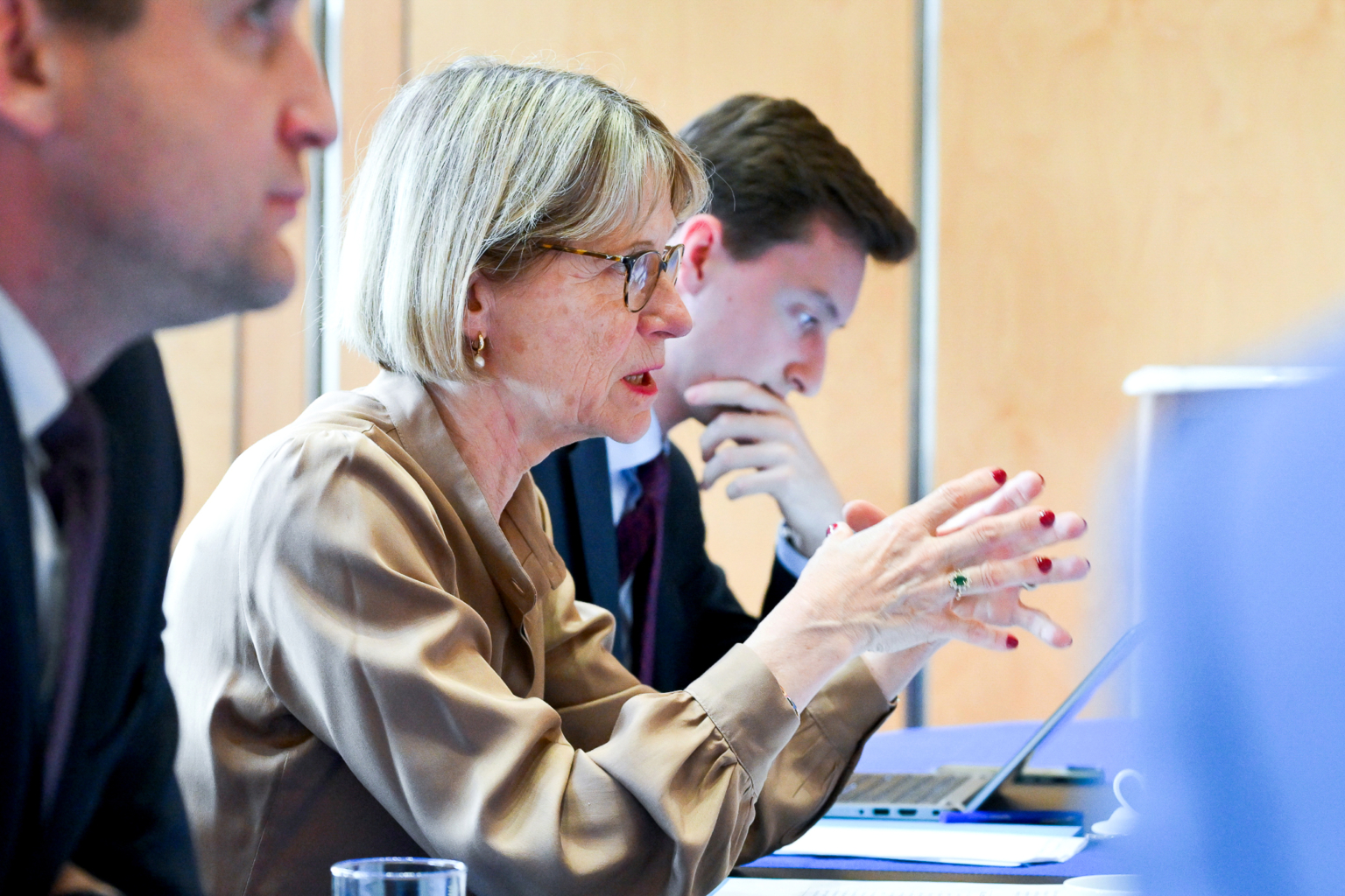 Une femme aux cheveux blonds courts et aux lunettes parle en faisant des gestes avec ses mains lors d'une réunion. Deux hommes en costume sont assis à côté d'elle, l'un d'eux se concentrant sur un ordinateur portable. Le cadre semble être celui d'un bureau professionnel ou d'une salle de conférence. Photographe Xavier Granet