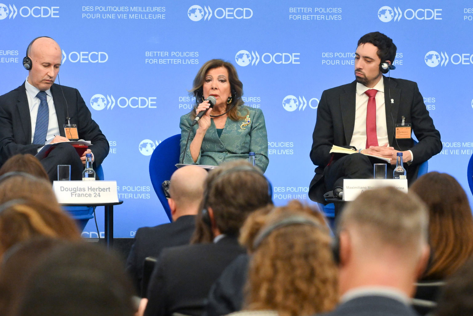 Trois panélistes sont assis sur la scène lors d'un événement de l'OCDE. La femme au centre parle dans un microphone tandis que les deux hommes à côté d'elle écoutent. L'arrière-plan affiche le logo de l'OCDE et le slogan "De meilleures politiques pour de meilleures vies". Photographe Xavier Granet