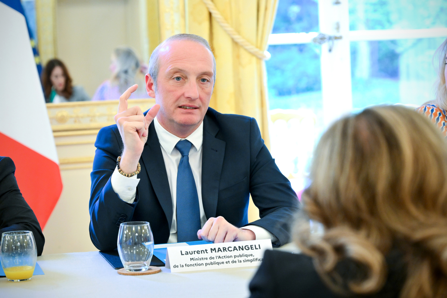 Un homme en costume, identifié par une plaque d'identification comme étant Laurent Marcangeli, parle et fait des gestes de la main lors d'une réunion. Deux personnes sont assises en face de lui à une table avec des boissons dans une pièce lumineuse et élégante. Photographe Xavier Granet