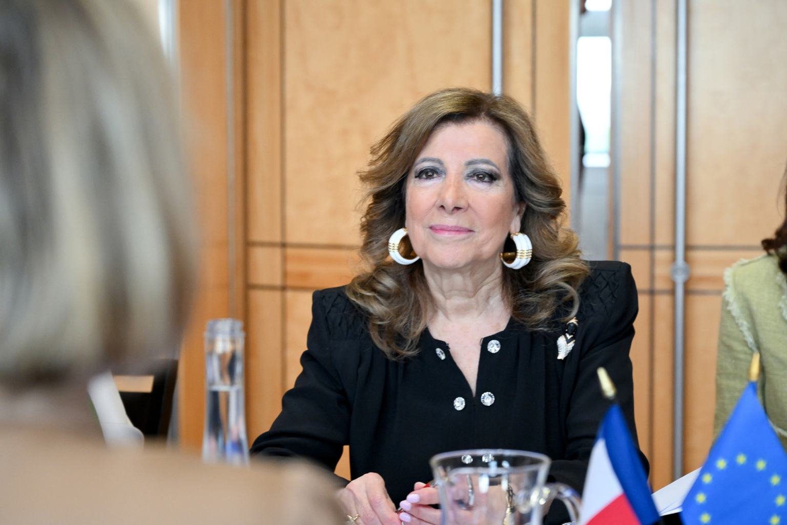 Une femme aux cheveux ondulés mi-longs et aux grandes boucles d'oreilles est assise à une table avec des drapeaux de la France et de l'Union européenne. Elle porte une veste noire et sourit légèrement. Une autre personne est floue au premier plan. Photographe Xavier Granet