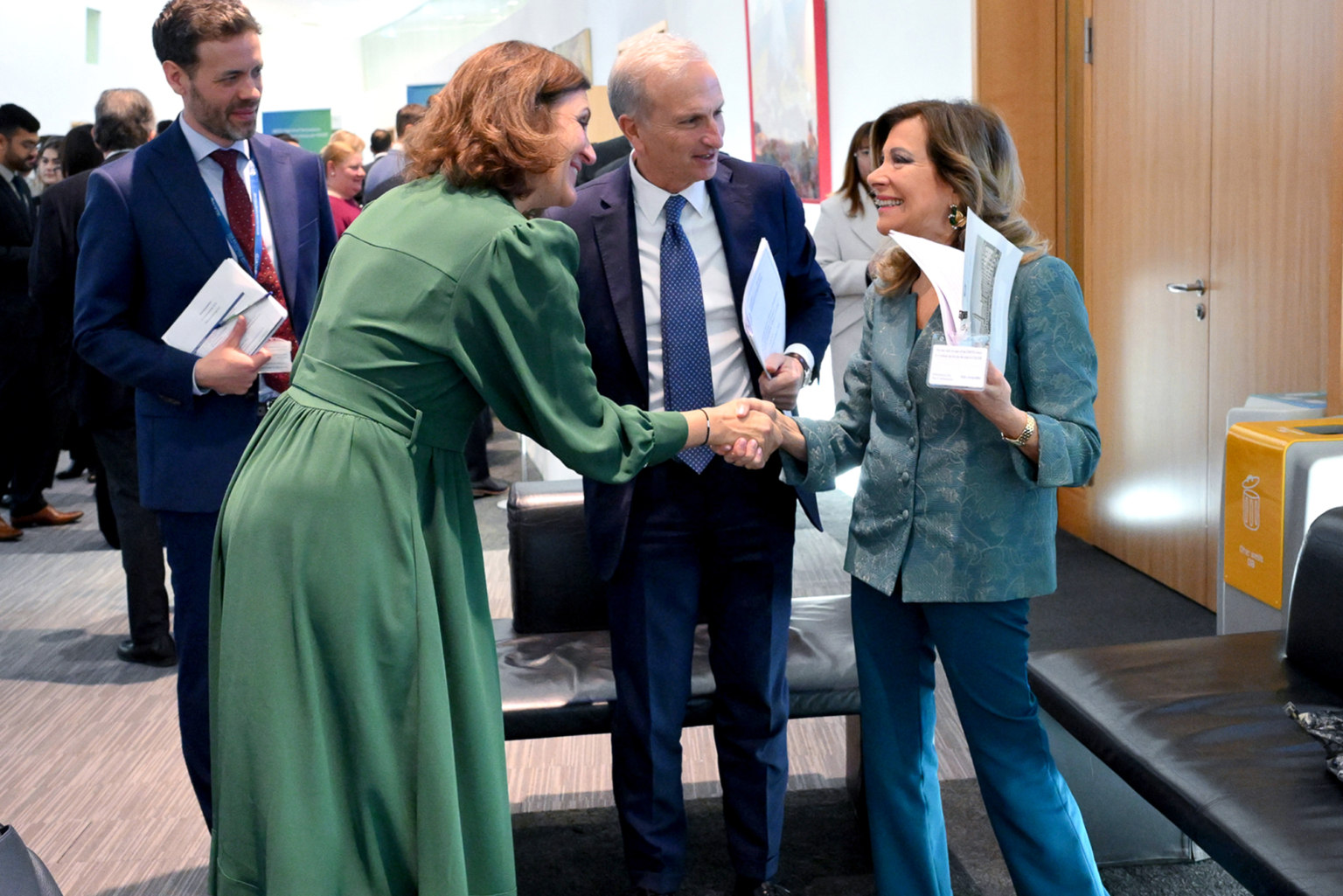 Quatre personnes habillées professionnellement se tiennent à l'intérieur lors d'un événement. Deux femmes au premier plan se serrent la main et sourient, tandis que deux hommes en costume se tiennent à proximité, observant et souriant. Il semble s'agir d'un bureau ou d'un espace de conférence. Photographe Xavier Granet