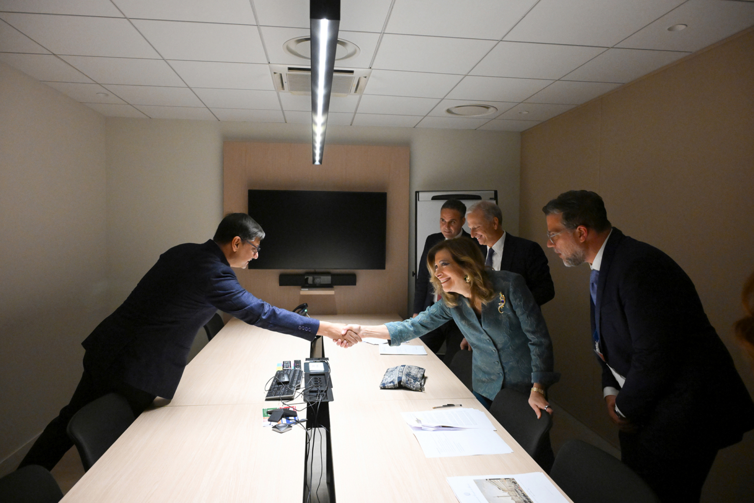 Cinq personnes se trouvent dans une salle de conférence. Deux d'entre elles, un homme et une femme, se penchent sur une longue table pour se serrer la main, tandis que trois autres se tiennent à proximité, observant et souriant. Un grand écran est fixé au mur derrière eux. Photographe Xavier Granet