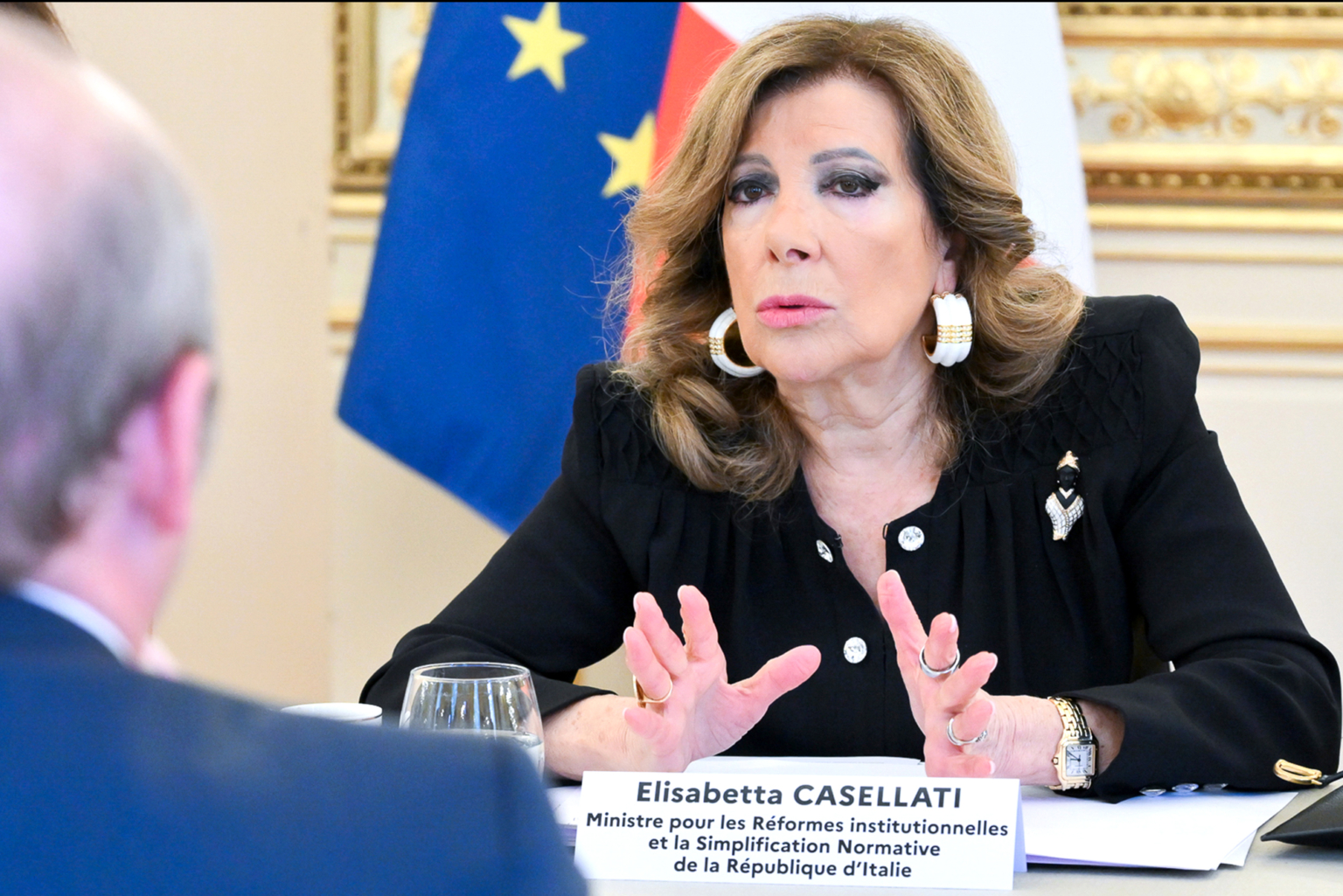 Une femme aux cheveux bruns ondulés, portant une tenue noire et de grandes boucles d'oreilles blanches, prend la parole lors d'une réunion. Une plaque devant elle indique "Elisabetta Casellati". Des drapeaux et des murs ornés sont visibles à l'arrière-plan. Photographe Xavier Granet