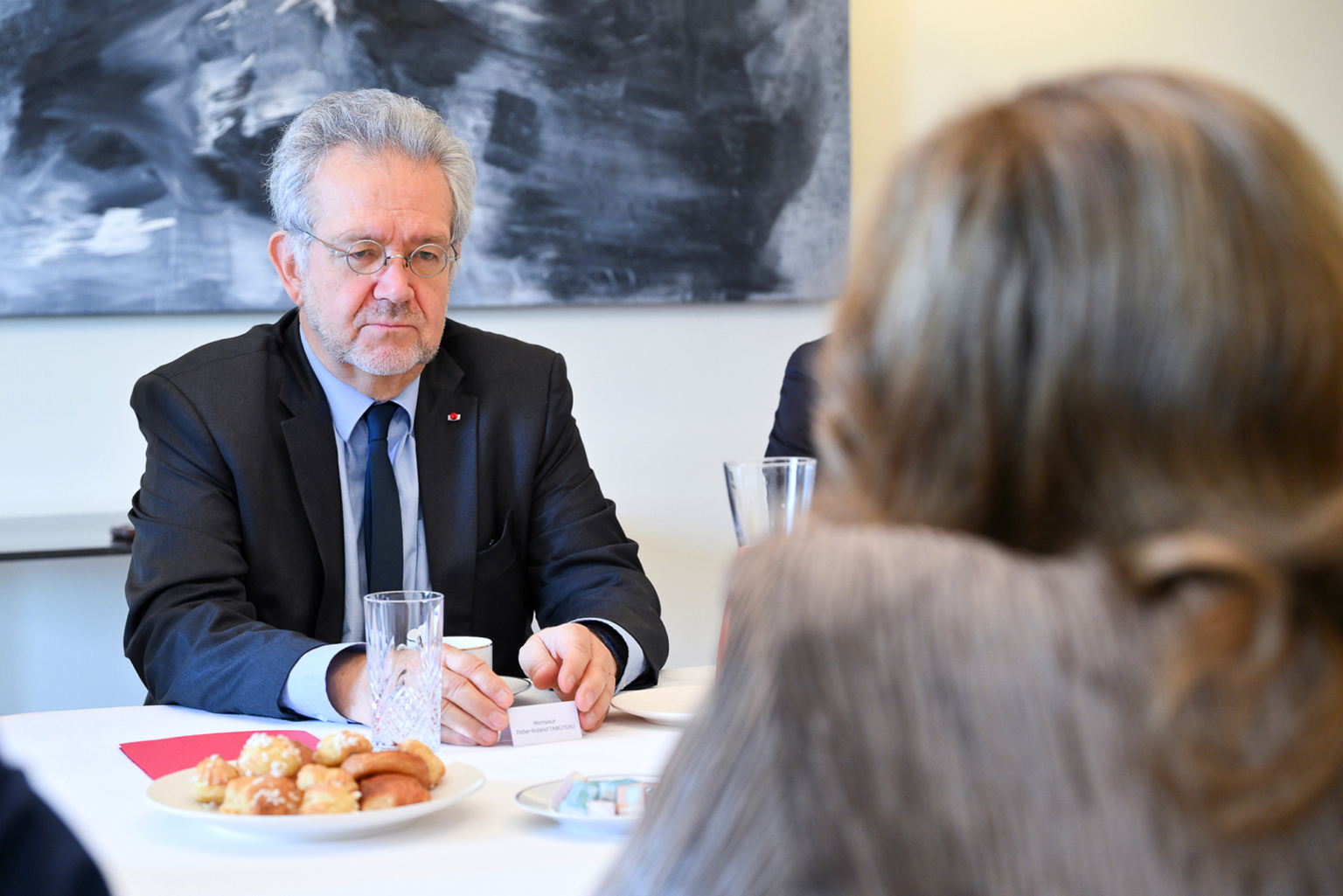 Un homme âgé en costume et lunettes est assis à une table avec des pâtisseries, un verre d'eau et une carte de notes, face à une personne aux cheveux longs. Le cadre semble être celui d'une réunion ou d'une discussion formelle. Photographe Xavier Granet