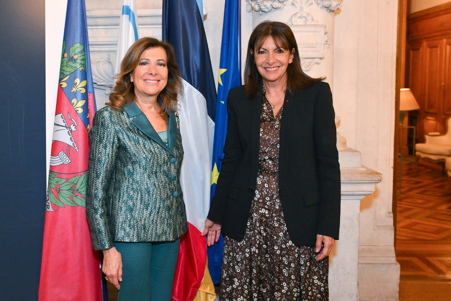 Deux femmes sourient à l'intérieur devant le drapeau français, le drapeau de l'Union européenne et un autre drapeau. Elles sont habillées de manière formelle, l'une en tailleur vert et l'autre en robe et veste sombres, dans une pièce ornée de détails en bois et en pierre. Photographe Xavier Granet