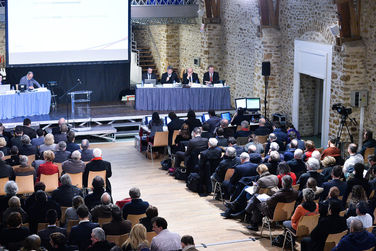 Un large public est assis face à un panel d'orateurs lors d'une conférence formelle dans une salle spacieuse aux murs en pierre et au sol en bois. Certaines personnes prennent des notes et des diapositives sont projetées sur le mur. Photographe Xavier Granet