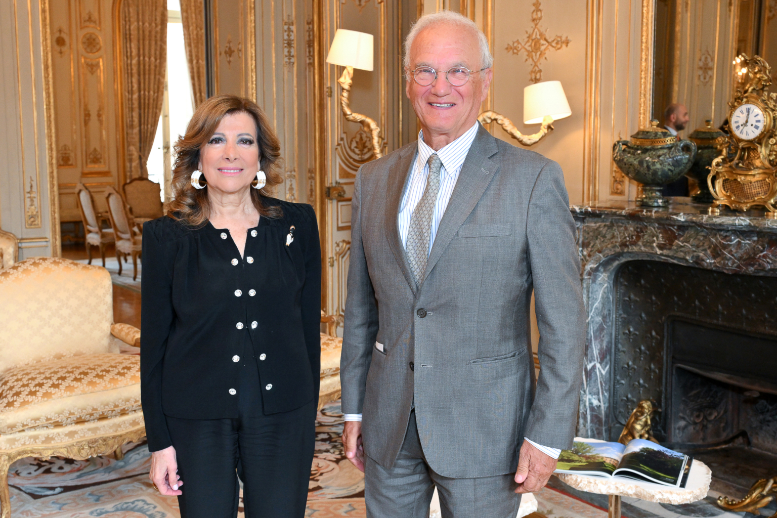 Une femme en tenue noire et un homme en costume gris sourient dans une pièce ornée de meubles élégants, d'une cheminée en marbre et d'un décor classique. Photographe Xavier Granet