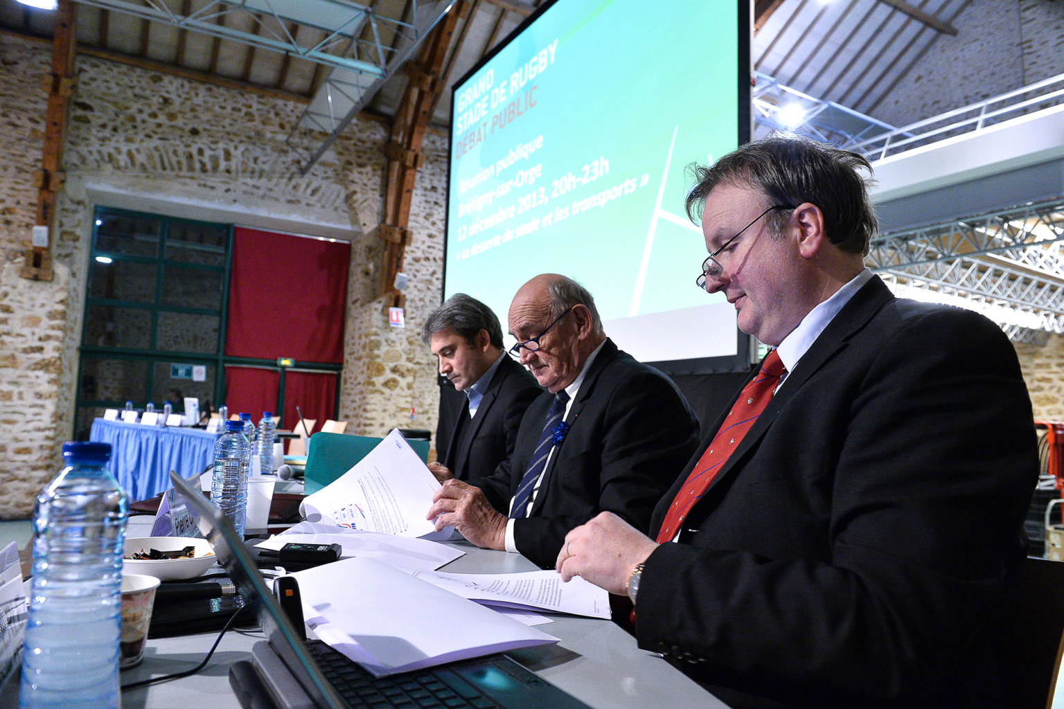 Trois hommes en costume sont assis à une table avec des documents et des ordinateurs portables dans une salle de conférence. Un grand écran avec du texte se trouve derrière eux, et des bouteilles d'eau et des snacks sont posés sur la table. La salle a des murs en pierre et de hauts plafonds. Photographe Xavier Granet