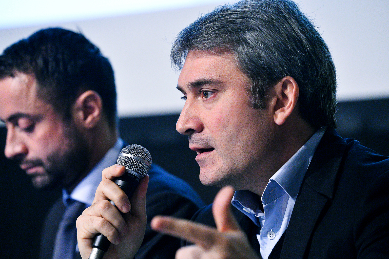 Deux hommes en costume sont assis côte à côte. L'homme au premier plan parle dans un microphone et fait des gestes de la main, tandis que l'homme à l'arrière-plan regarde vers le bas. Tous deux semblent participer à un événement ou à une discussion formelle. Photographe Xavier Granet