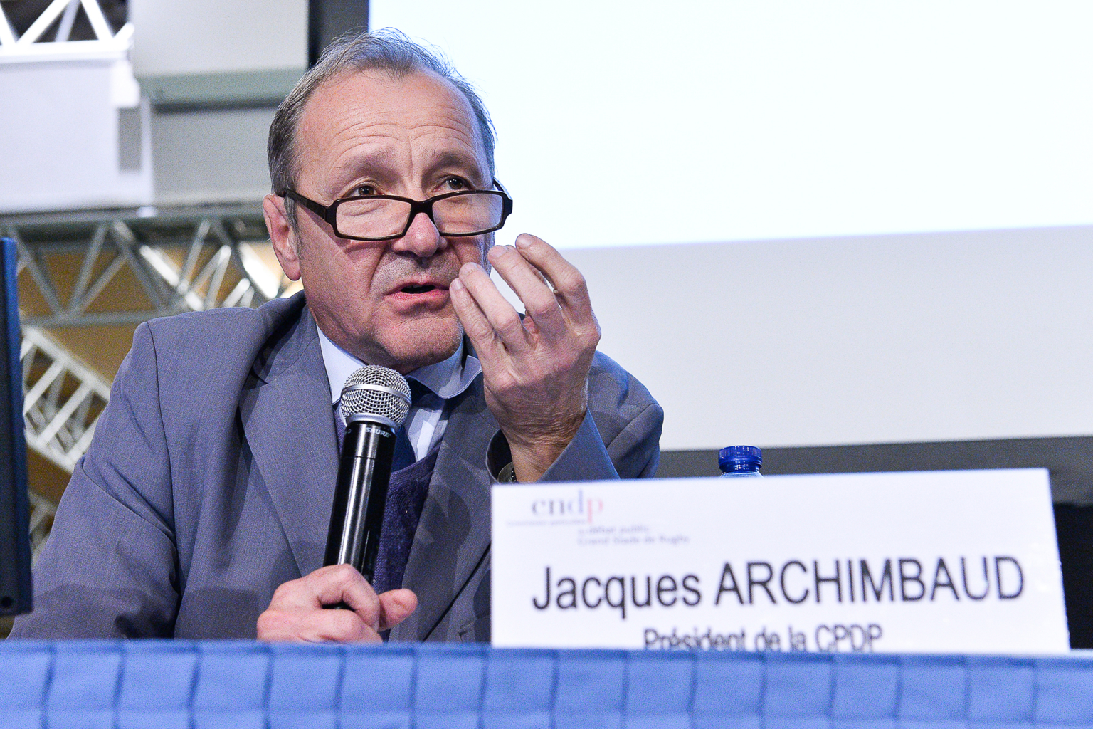 Un homme en costume et lunettes parle dans un micro et tient un objet à la main à une table de conférence. Devant lui, une plaque indique "Jacques Archimbaud, Président de la CPDP". Photographe Xavier Granet