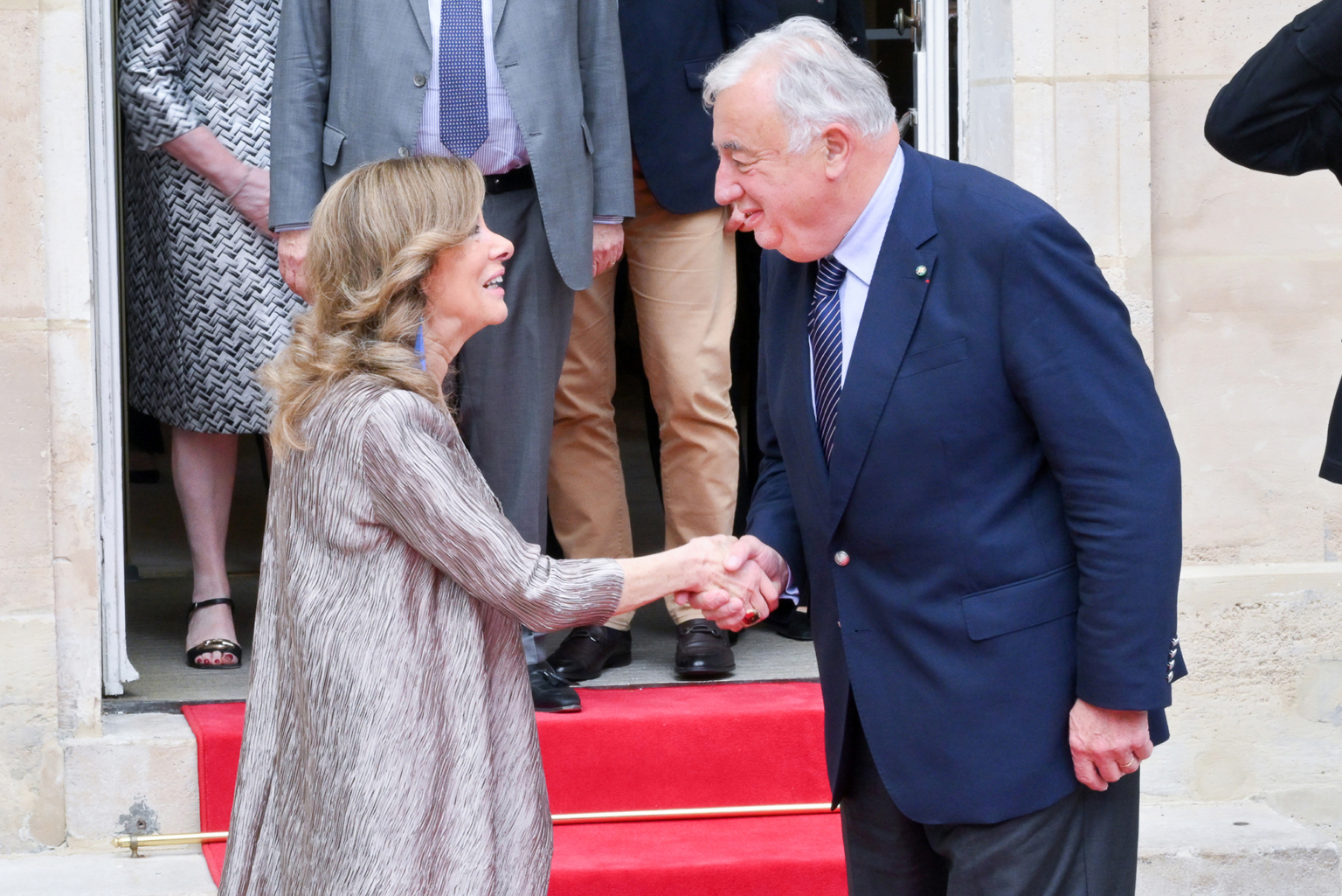 Une femme et un homme plus âgé se serrent la main et se sourient sur un tapis rouge à l'extérieur d'un bâtiment, plusieurs personnes se tenant à l'arrière-plan près de la porte. Photographe Xavier Granet