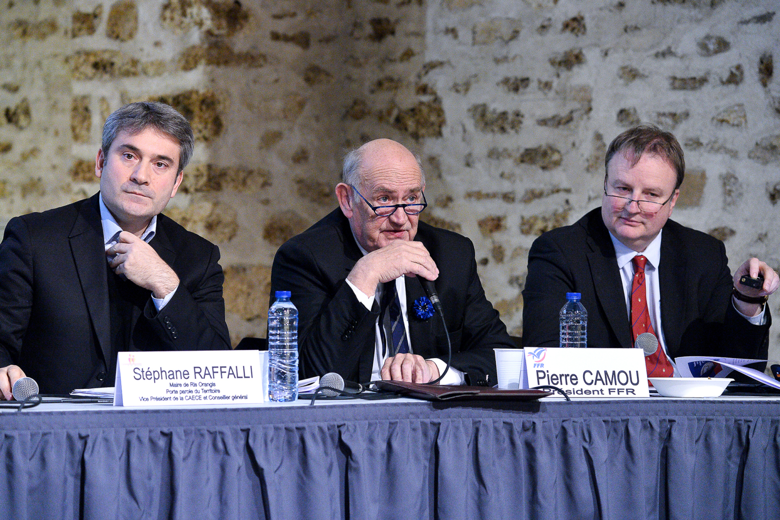 Trois hommes en costume sont assis à une table avec des microphones et des plaques nominatives devant eux, prenant la parole lors d'une conférence ou d'une réunion. Des bouteilles d'eau et des papiers sont posés sur la table, et un mur de pierre se trouve à l'arrière-plan. Photographe Xavier Granet