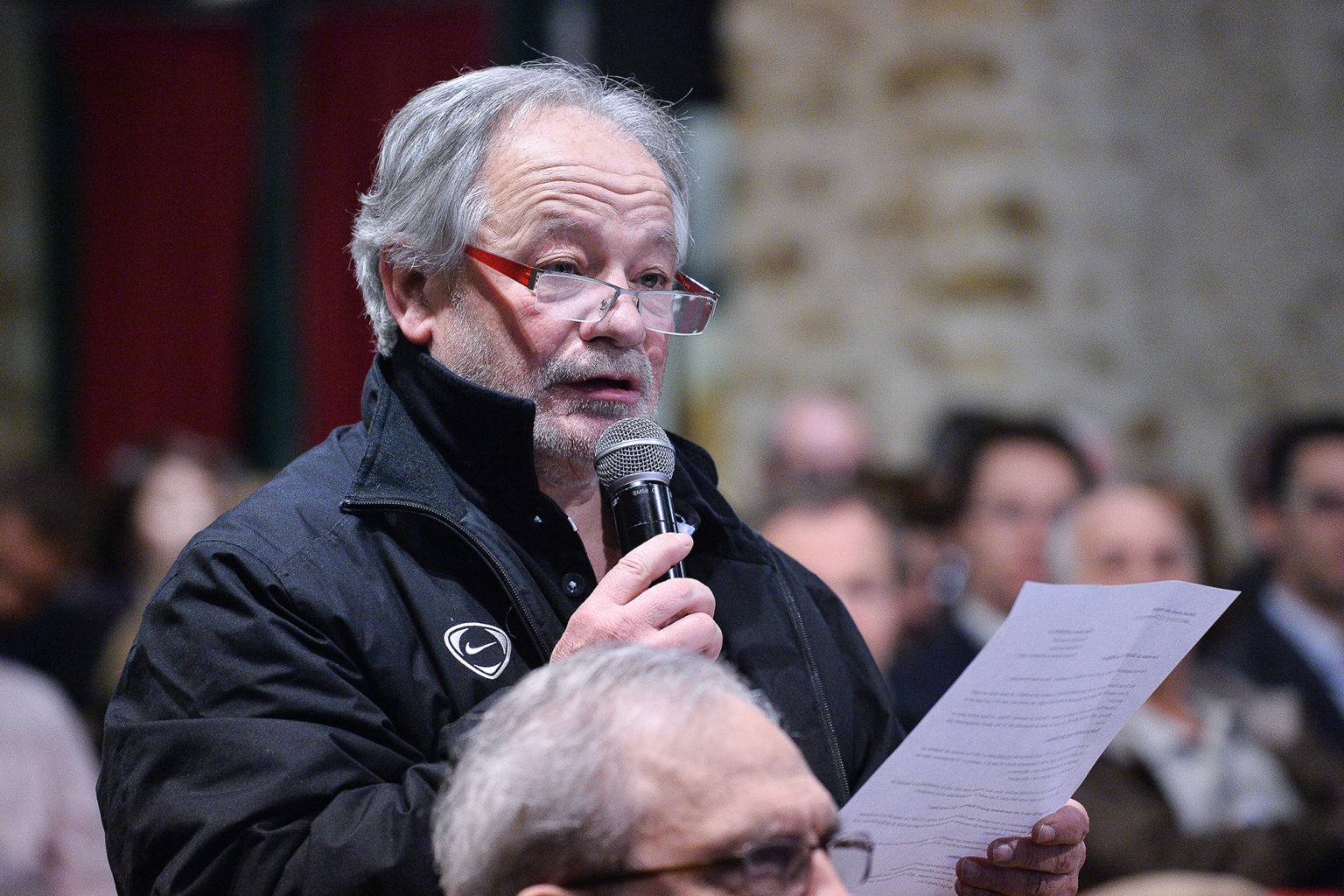 Un homme âgé aux cheveux gris et aux lunettes tient un microphone et une feuille de papier pendant qu'il parle lors d'un événement en intérieur. Des personnes sont assises dans l'arrière-plan flou. Photographe Xavier Granet
