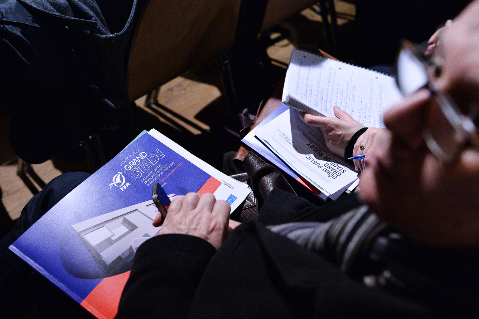 Une personne tenant un livret "Grand Stade" et un stylo est assise à côté d'une personne prenant des notes dans un carnet lors d'une réunion ou d'une présentation. Les deux personnes sont concentrées sur leurs documents. Photographe Xavier Granet