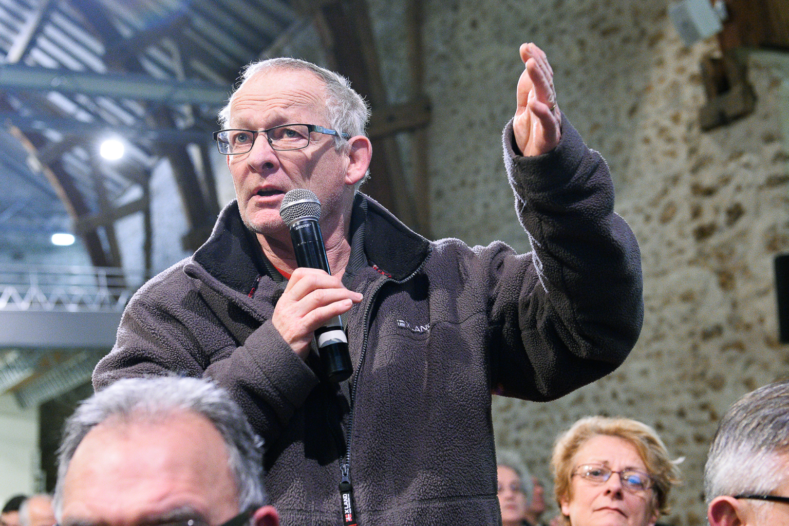 Un homme âgé portant des lunettes et une veste polaire parle dans un microphone en levant une main, debout parmi un public assis dans un décor intérieur rustique avec des murs en pierre. Photographe Xavier Granet