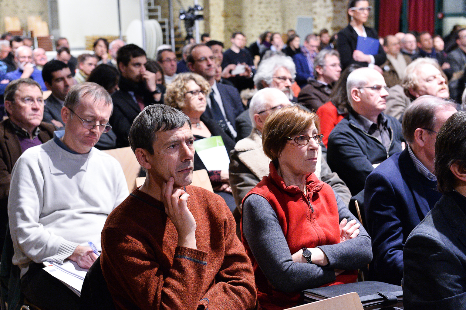 Un grand groupe de personnes est assis dans une salle bien éclairée, écoutant attentivement un orateur hors champ. Certains prennent des notes, tandis que d'autres regardent ou s'assoient en croisant les bras. Le public semble concentré et engagé. Photographe Xavier Granet