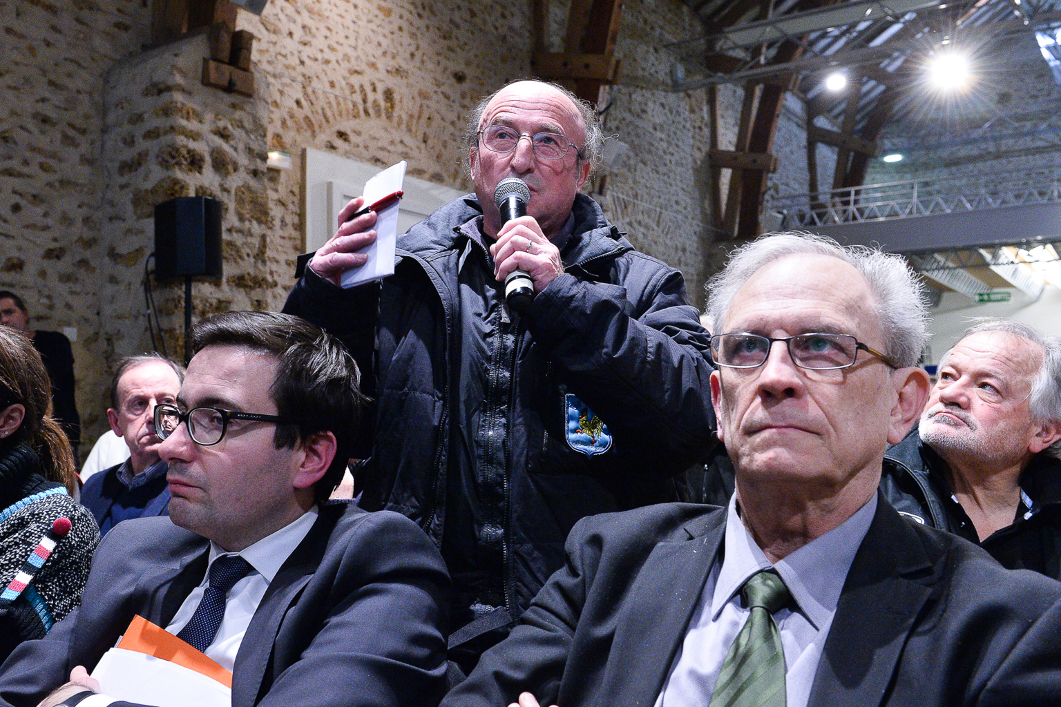 Un homme d'un certain âge, portant des lunettes et une veste sombre, parle dans un microphone au milieu de participants assis lors d'un événement en intérieur. D'autres hommes en costume et lunettes sont assis au premier plan, écoutant attentivement. Photographe Xavier Granet