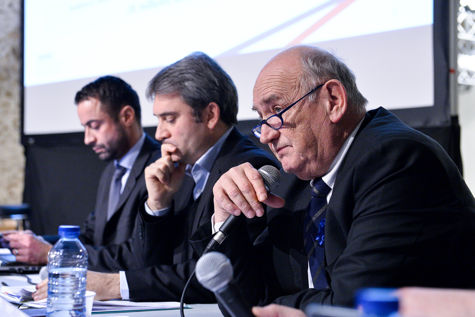 Trois hommes en costume sont assis à une table lors d'une conférence ou d'une table ronde. L'homme au premier plan parle dans un microphone tandis que les autres écoutent et prennent des notes. Des bouteilles d'eau et des papiers sont posés sur la table devant eux. Photographe Xavier Granet