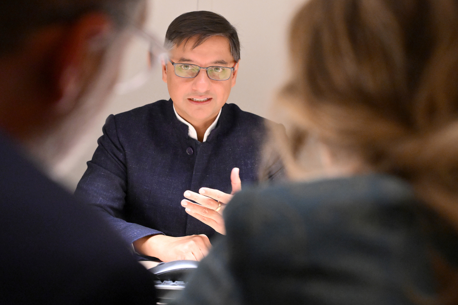 Un homme portant des lunettes et un blazer sombre est assis à une table et fait un geste de la main tout en parlant à deux personnes dont on voit le dos au premier plan. Photographe Xavier Granet