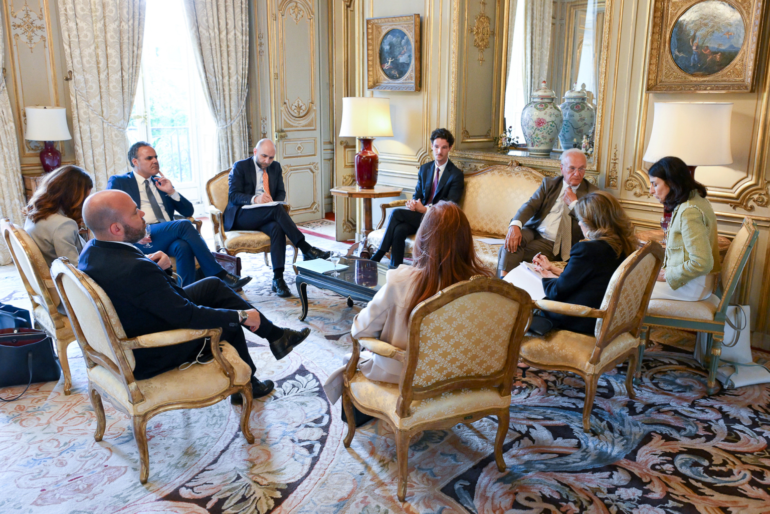 Un groupe de neuf personnes est assis sur des chaises ornées autour d'une table basse en verre, dans une pièce élégante et décorée de dorures, avec de grandes fenêtres, des vases décoratifs et des œuvres d'art encadrées, et participe à une discussion formelle. Photographe Xavier Granet