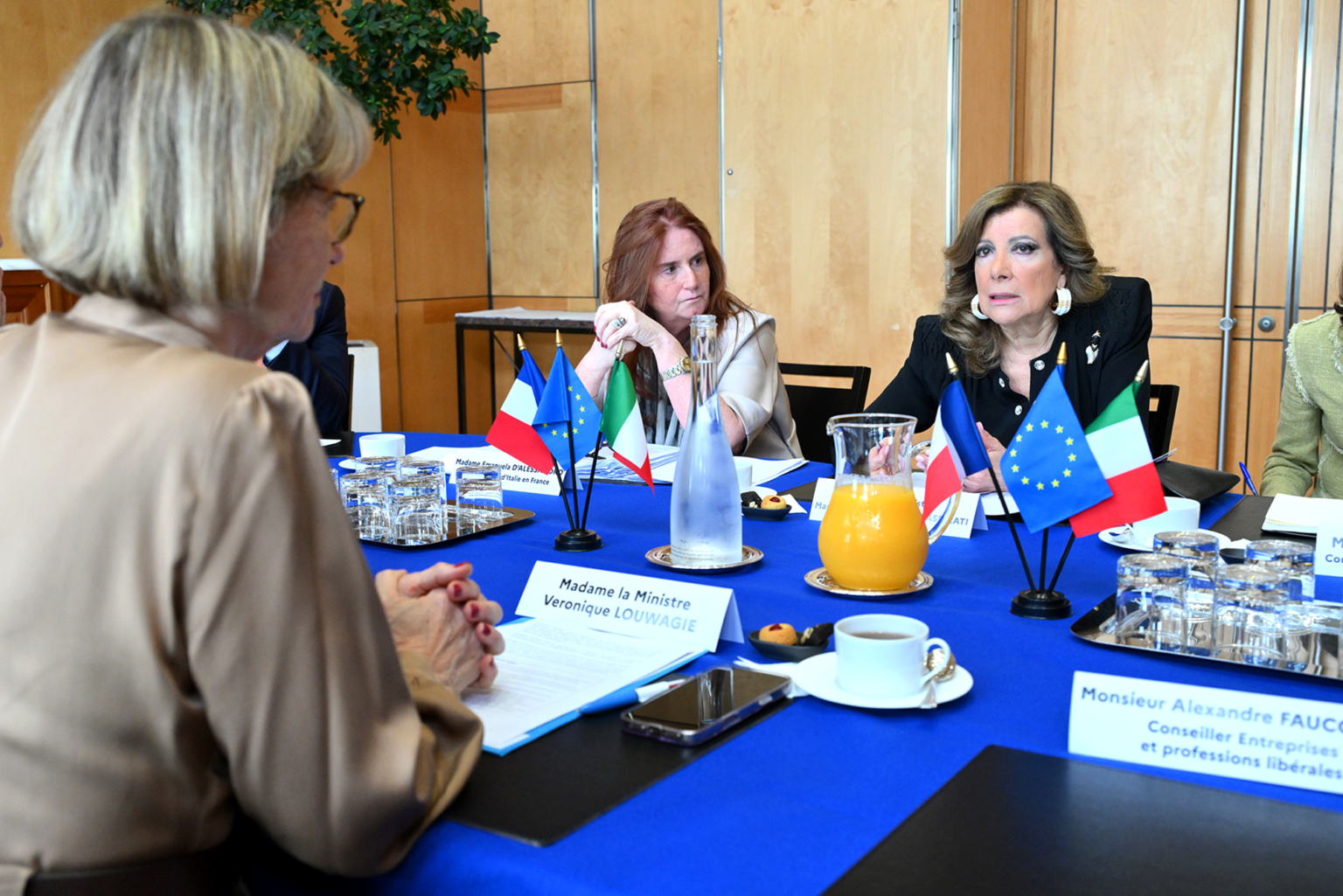 Trois femmes sont assises à une table de conférence avec des drapeaux européens et nationaux, des documents et des boissons. L'atmosphère semble formelle et sérieuse, une femme parlant tandis que les autres écoutent attentivement. Photographe Xavier Granet