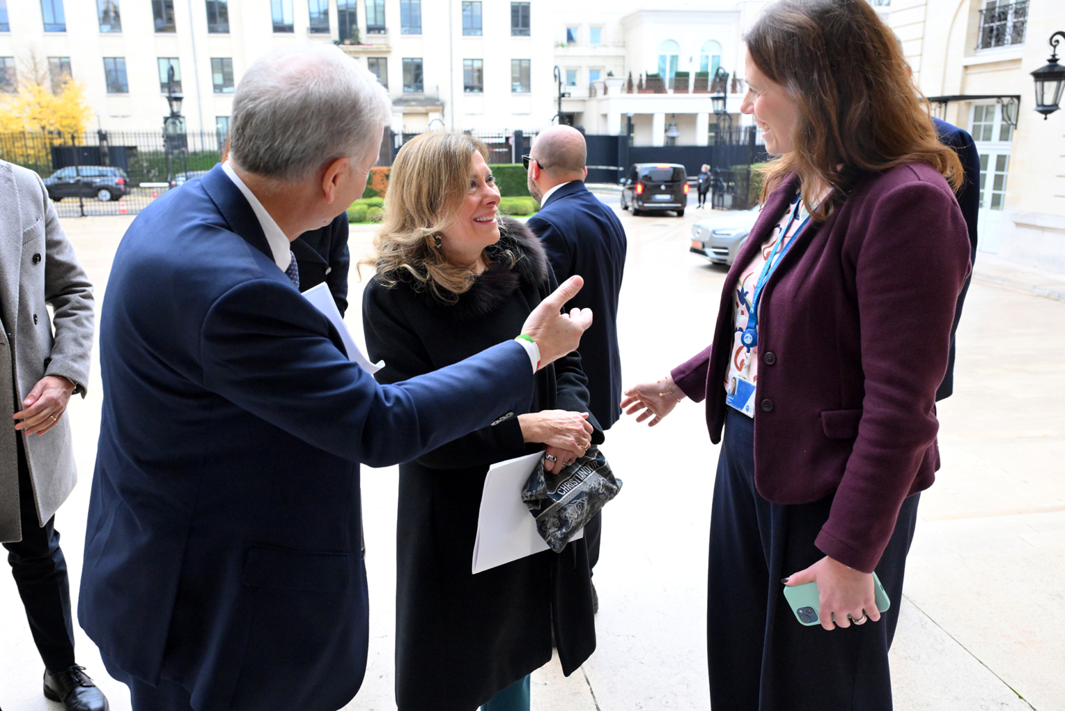 Un homme et deux femmes se tiennent à l'extérieur d'un bâtiment, souriant et discutant. L'une des femmes tient des papiers et un petit sac, l'autre un téléphone. Des personnes et des voitures se trouvent à l'arrière-plan, près d'un bâtiment moderne. Photographe Xavier Granet