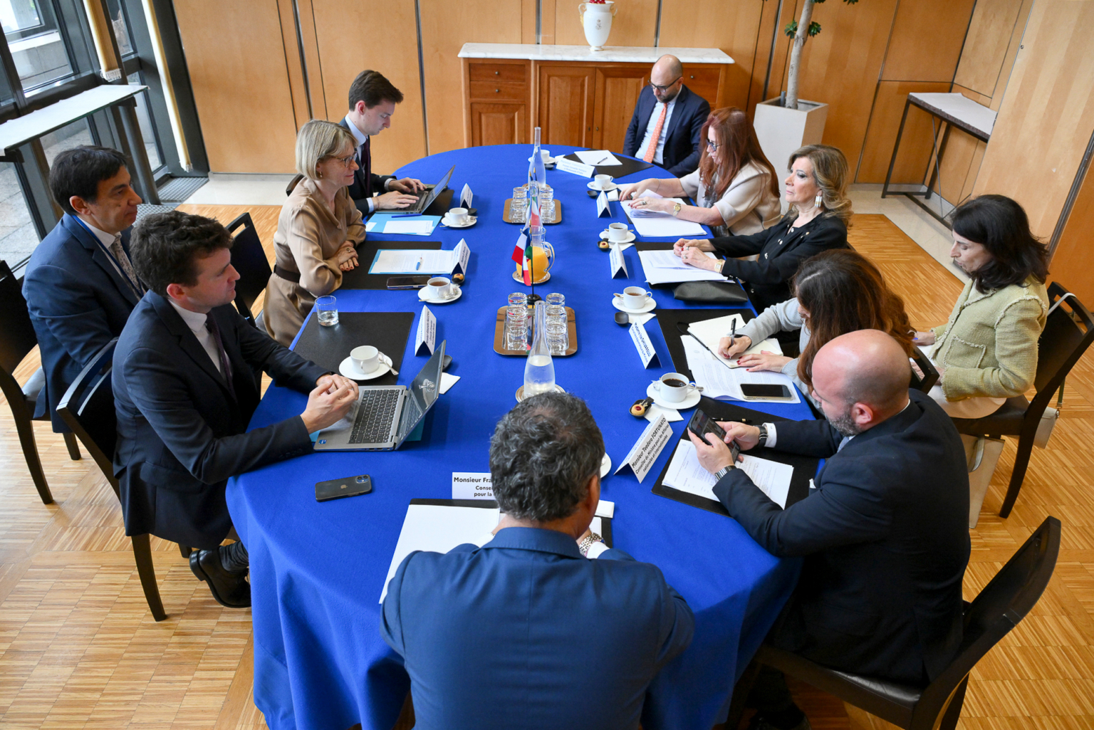 Un groupe de personnes en tenue de travail est assis autour d'une table recouverte d'une nappe bleue dans une salle de réunion, discutant de documents et utilisant des ordinateurs portables. Des drapeaux et des tasses de café sont posés sur la table, et une armoire se trouve à l'arrière-plan. Photographe Xavier Granet