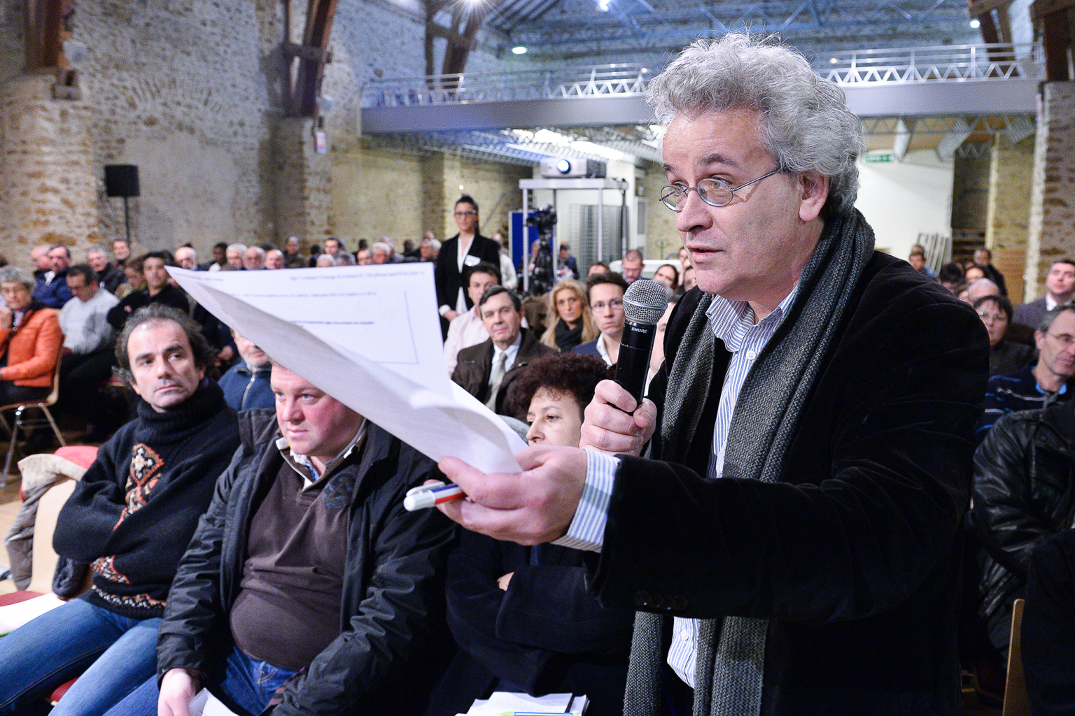 Un homme tenant des documents et un microphone s'adresse à un public assis dans une grande salle aux murs de pierre. Les gens écoutent attentivement et l'atmosphère semble être celle d'une réunion publique ou d'un événement communautaire. Photographe Xavier Granet