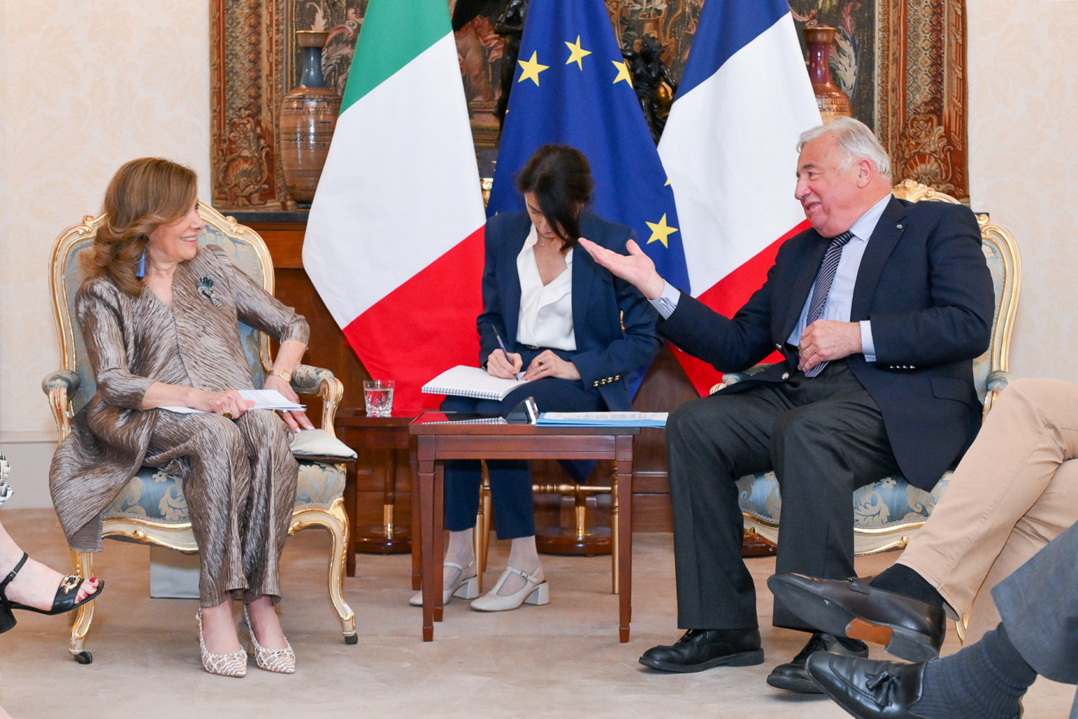 Trois personnes sont assises sur des chaises ornées, en pleine conversation, avec des drapeaux italiens et français derrière elles. Deux d'entre elles parlent et sourient, tandis que la troisième prend des notes. Le cadre semble formel et diplomatique. Photographe Xavier Granet
