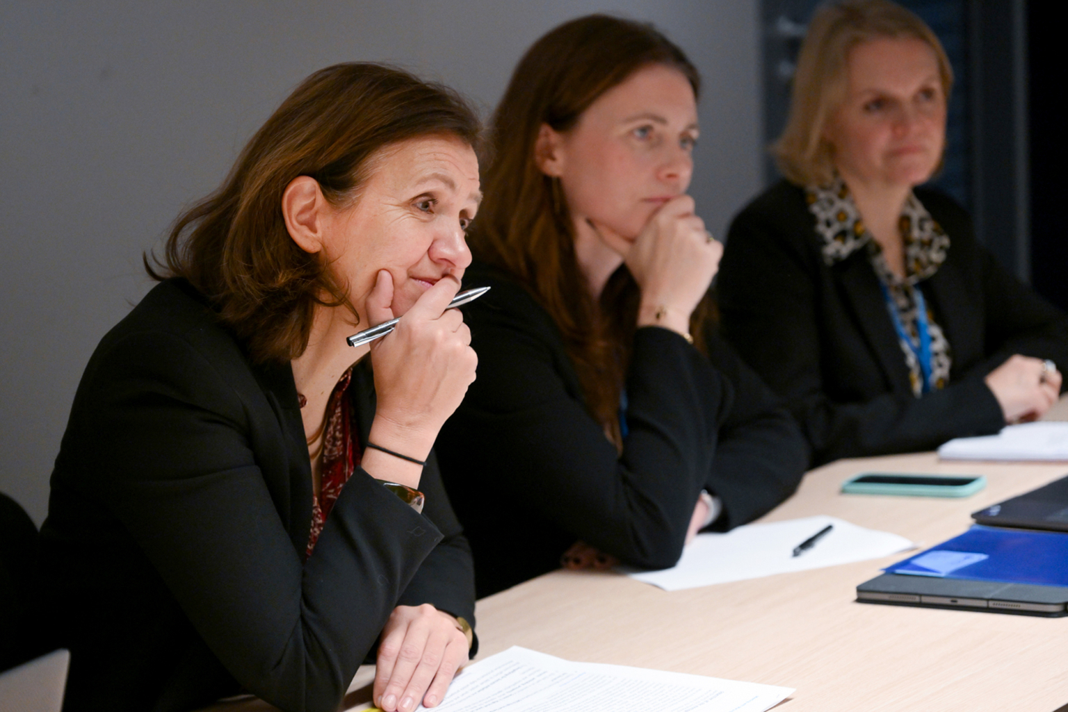 Trois femmes en tenue de travail sont assises à une table, l'air attentif et pensif. L'une d'elles porte un stylo à son menton, et des papiers et des carnets sont posés sur la table devant elles. Elles semblent engagées dans une réunion ou une discussion. Photographe Xavier Granet