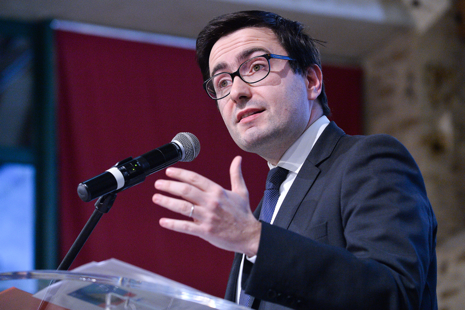 Un homme en costume et lunettes parle dans un micro sur un podium, en faisant un geste de la main. Une toile de fond rouge et un mur de pierre sont visibles à l'arrière-plan. Photographe Xavier Granet