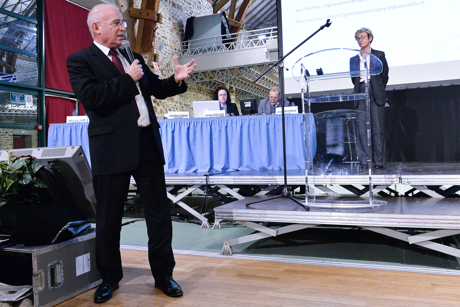 Un homme en costume parle dans un micro, debout sur un plancher en bois devant une scène, où trois personnes sont assises à une table avec des ordinateurs portables et une autre personne se tient debout sur un podium clair. Photographe Xavier Granet