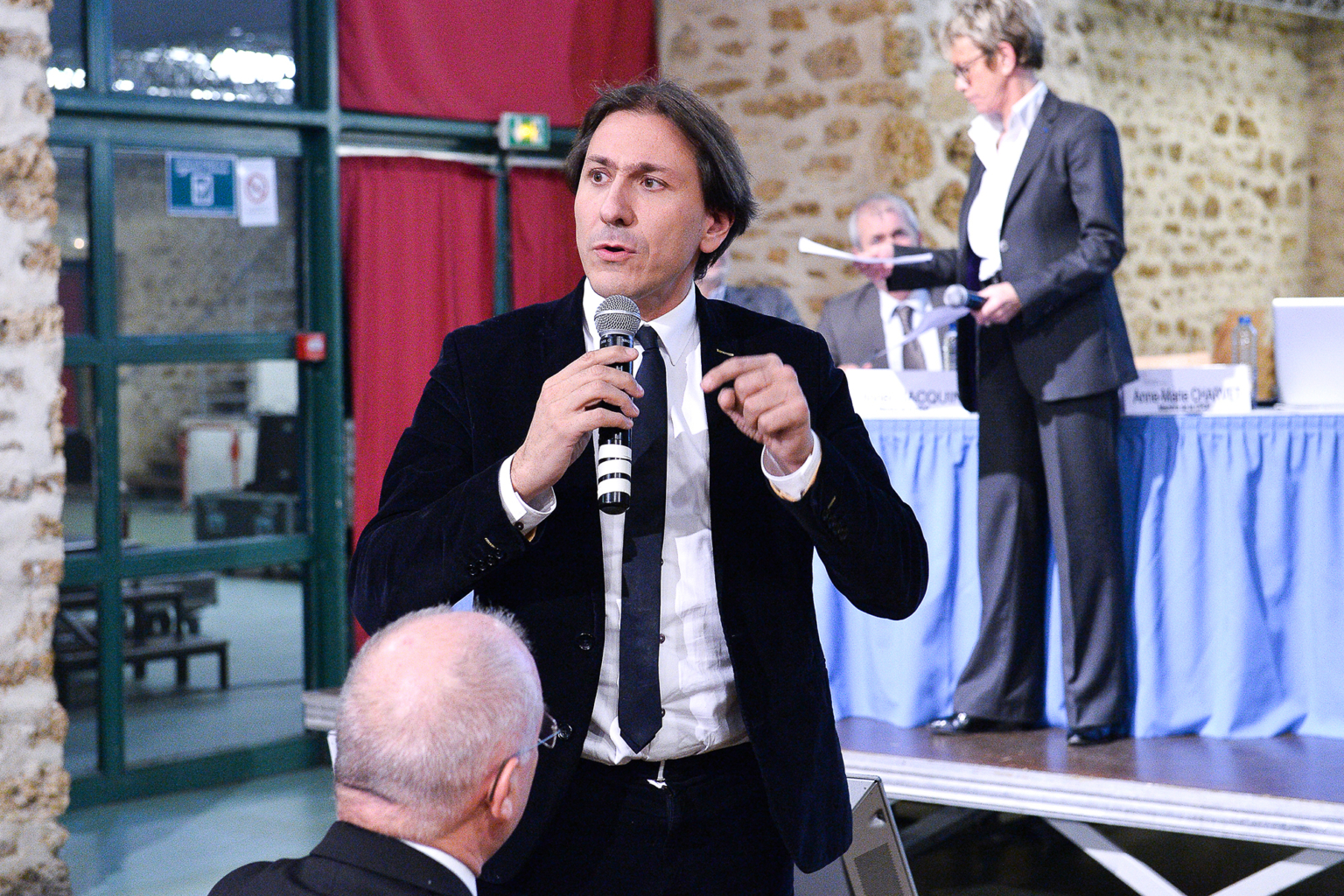 Un homme en costume sombre parle dans un microphone lors d'un événement en intérieur, tandis que des personnes sont assises à proximité et qu'un groupe de fonctionnaires est assis à une table drapée de bleu sur la scène à l'arrière-plan. Photographe Xavier Granet