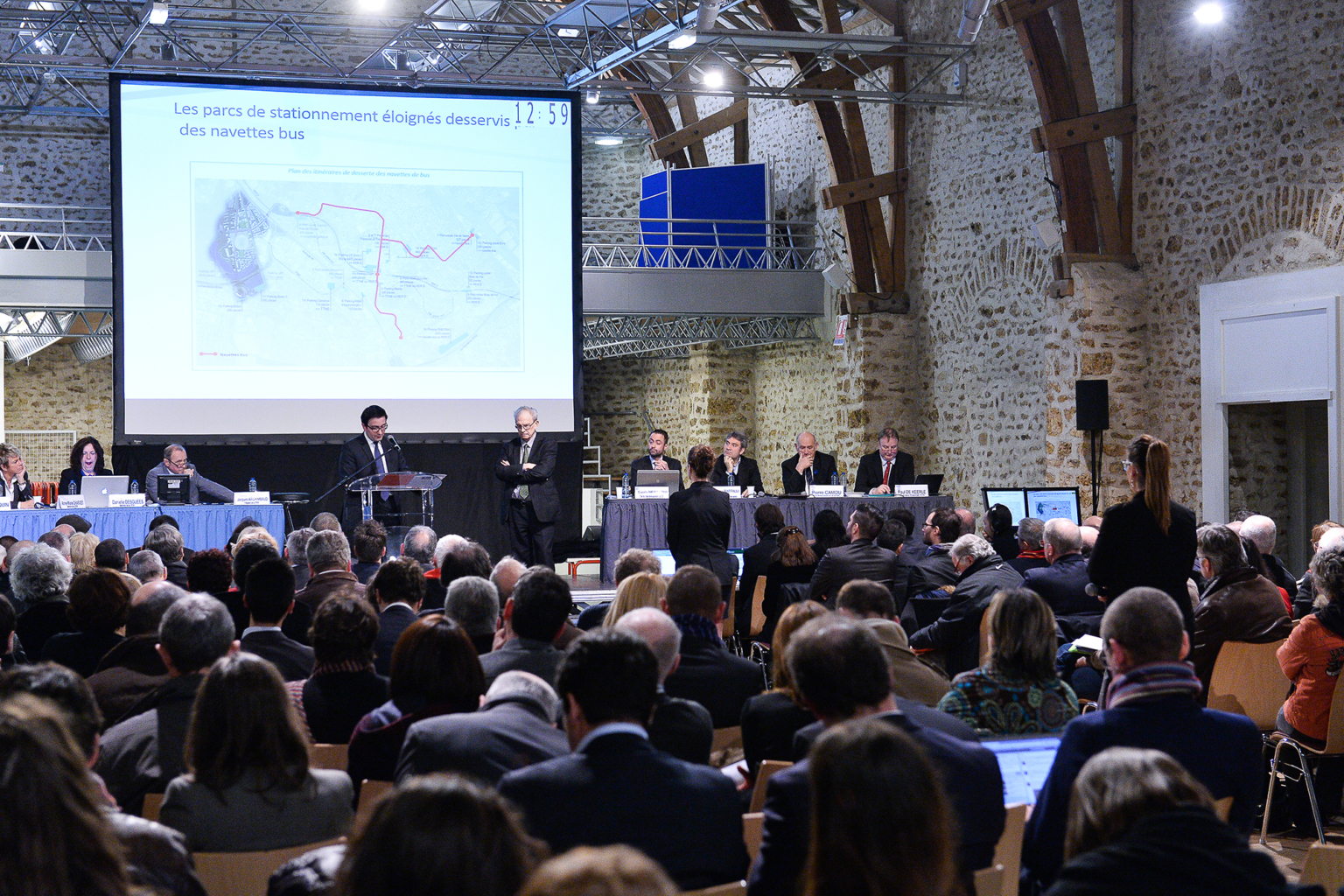 Un large public assiste à une présentation formelle dans une salle de conférence. Un orateur se tient sur un podium et une carte avec un texte en français est projetée sur un écran. Plusieurs personnes sont assises à des tables qui font face au public. Photographe Xavier Granet